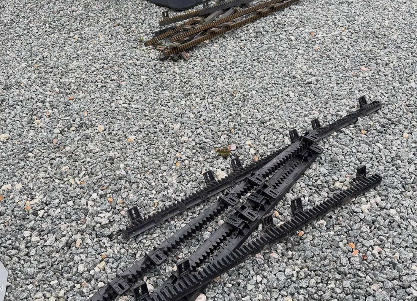 Black nylon gear rack sections for a sliding gate opener laid out on a gravel drive