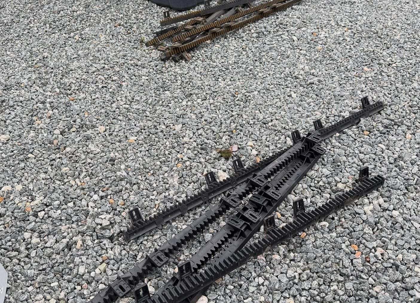 Black nylon gear rack sections for a sliding gate opener laid out on a gravel drive