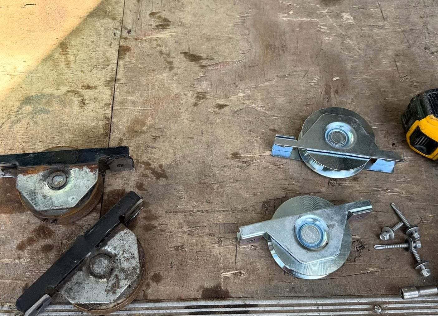 Two worn, rusted sliding gate rollers beside two new galvanized rollers and their bolts on a plywood bench