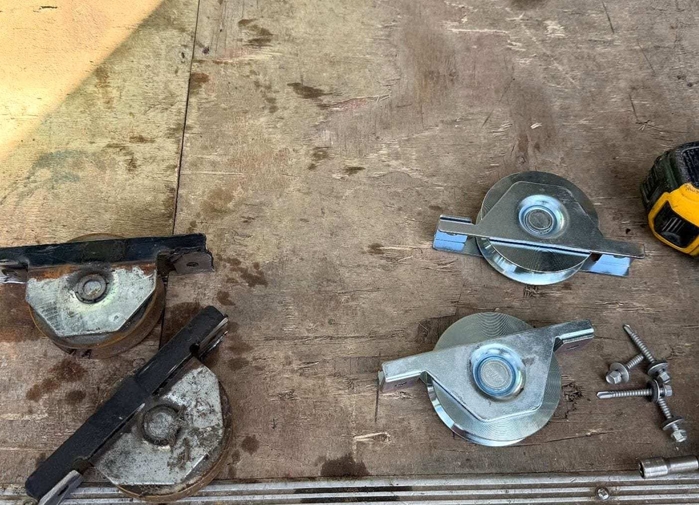 Two worn, rusted sliding gate rollers beside two new galvanized rollers and their bolts on a plywood bench