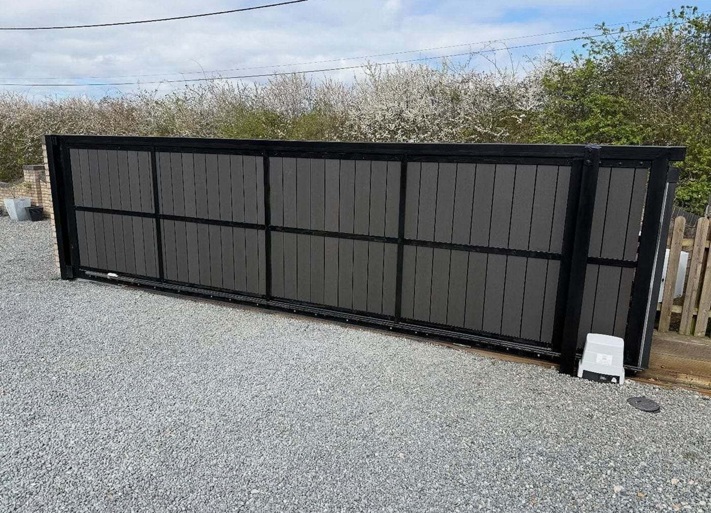 Long black sliding gate with dark composite panels across a gravel driveway, its opener at the end of the track