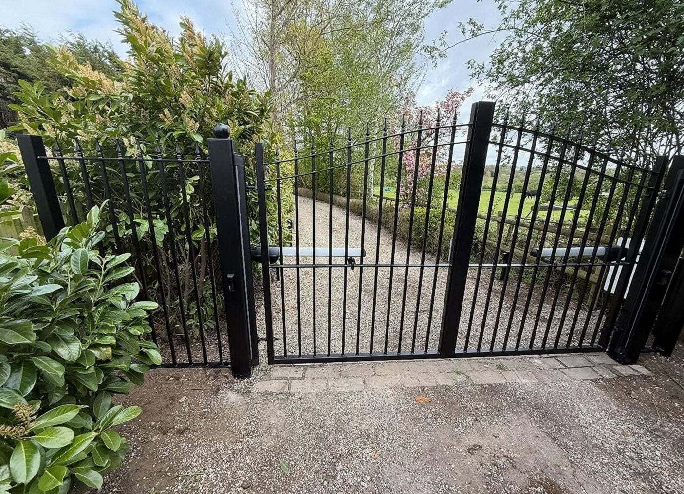 Black metal double swing gates with an arched top and a matching walk-through gate beside a garden path