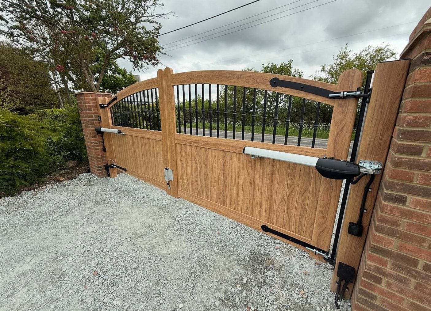 Wood-grain double swing gates with an arched railing top, hung on brick piers with operator arms on the inside